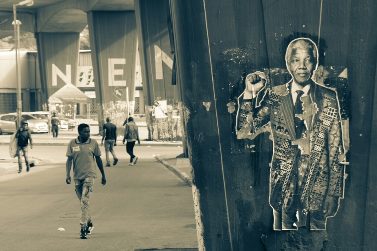 A weathered portrait of Nelson Mandela raising his fist is pasted to a concrete pillar beneath an overpass, while pedestrians walk along the street in the background.