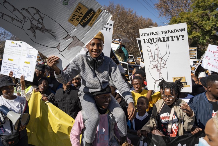 A smiling young protester sits on another participant’s shoulders while demonstrators march with placards calling for justice, freedom, and equality during a Youth Day protest in Pretoria.