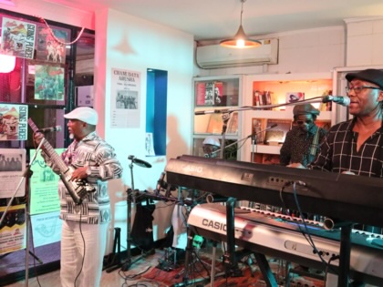 Members of the Kenyan band Them Mushrooms perform at the Santuri Salon in Nairobi, with keyboards, guitars, microphones, and recording equipment in a small venue lined with music posters and shelves of books and records.