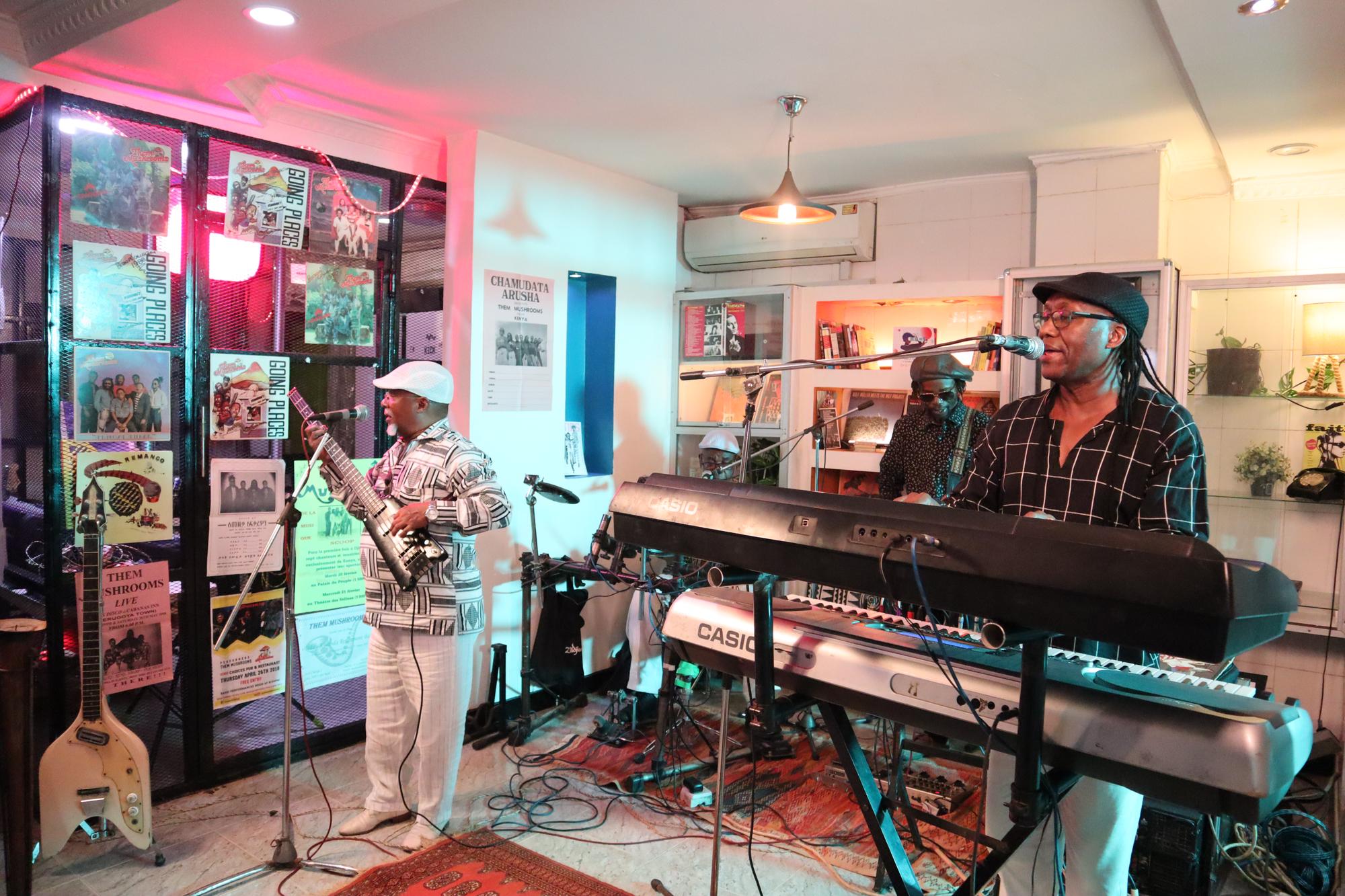 Members of the Kenyan band Them Mushrooms perform at the Santuri Salon in Nairobi, with keyboards, guitars, microphones, and recording equipment in a small venue lined with music posters and shelves of books and records.