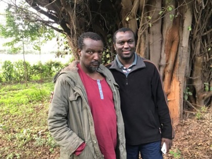 Two men stand in a forest in front of a large tree.