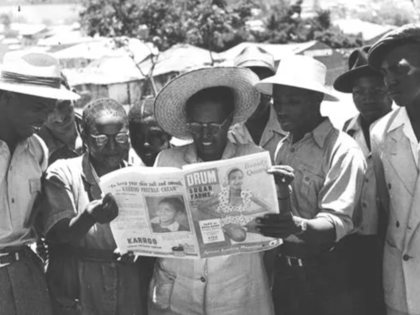 A group of Black South Africans gather outdoors around a man holding a copy of Drum magazine’s February 1953 issue, with several people leaning in to read the cover together. 