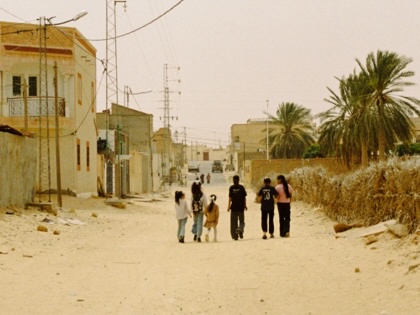 A group of children and young people walk down a sandy, unpaved road lined with low-rise homes, palm trees, and power lines in a residential neighborhood in Tunisia.
