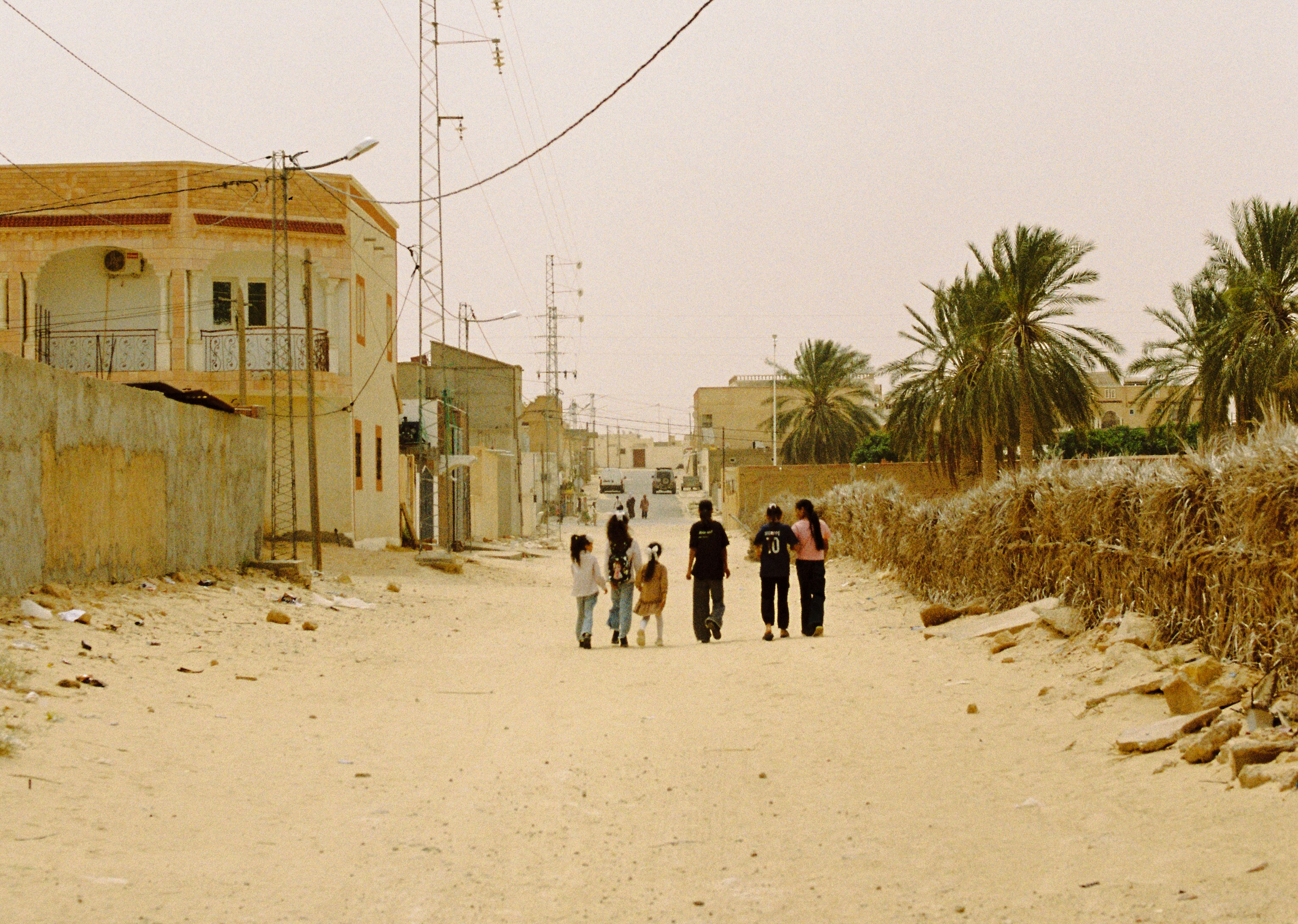 A group of children and young people walk down a sandy, unpaved road lined with low-rise homes, palm trees, and power lines in a residential neighborhood in Tunisia.