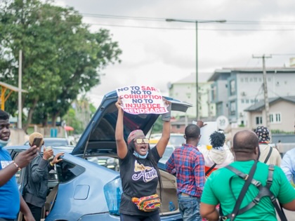 A protester raises a “#EndSARS” placard above a crowd gathered beside parked cars during a demonstration in Lagos, Nigeria, while photographers and other participants stand nearby.