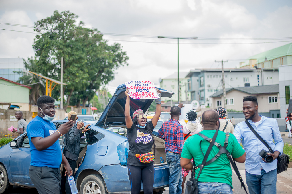 A protester raises a “#EndSARS” placard above a crowd gathered beside parked cars during a demonstration in Lagos, Nigeria, while photographers and other participants stand nearby.