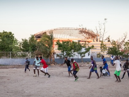 A group of boys play football on a dusty pitch in Beira, Mozambique, with the damaged roof of Macuti Cathedral visible in the background.
