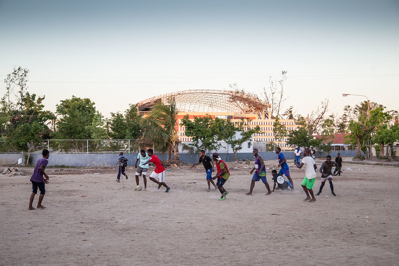 A group of boys play football on a dusty pitch in Beira, Mozambique, with the damaged roof of Macuti Cathedral visible in the background.