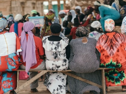 A group of women in colorful traditional dress sit on a bench in a community meeting in Ghana.