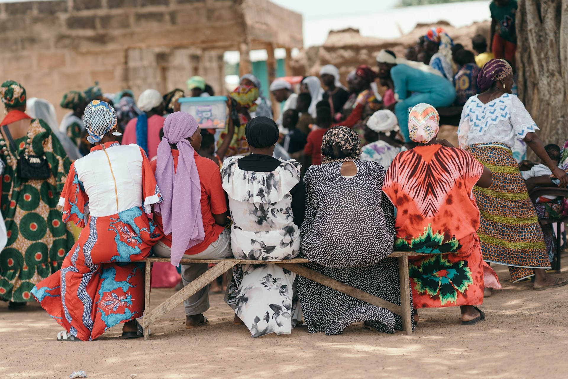 A group of women in colorful traditional dress sit on a bench in a community meeting in Ghana.