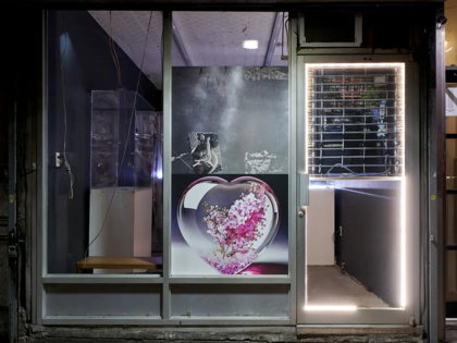 Nighttime exterior of Blade Study gallery in New York during YATTA’s FATHAGOD.EXE, with a large image of pink flowers inside a glass heart displayed in the storefront and a brightly lit entrance beside it.