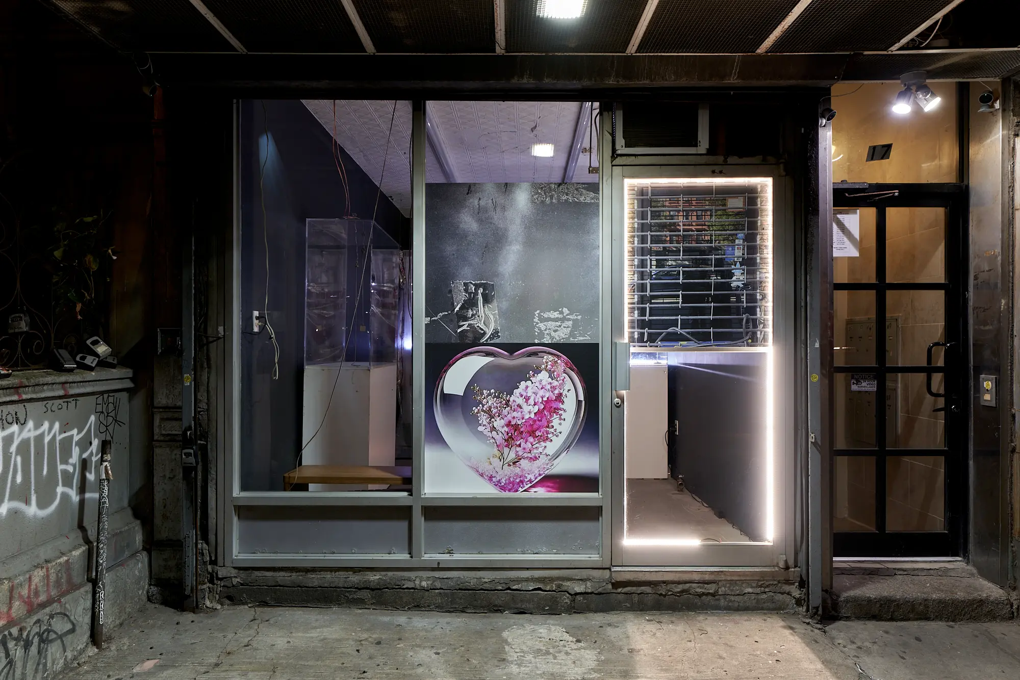 Nighttime exterior of Blade Study gallery in New York during YATTA’s FATHAGOD.EXE, with a large image of pink flowers inside a glass heart displayed in the storefront and a brightly lit entrance beside it.