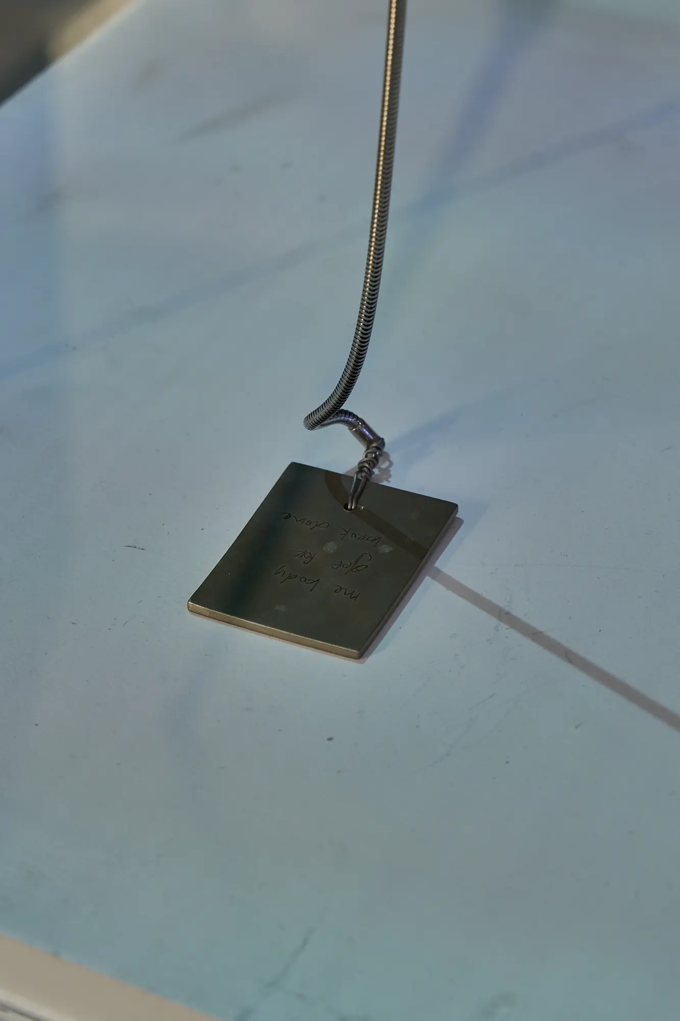 A close-up of an engraved metal pendant on a chain, resting on a pale marbled surface.
