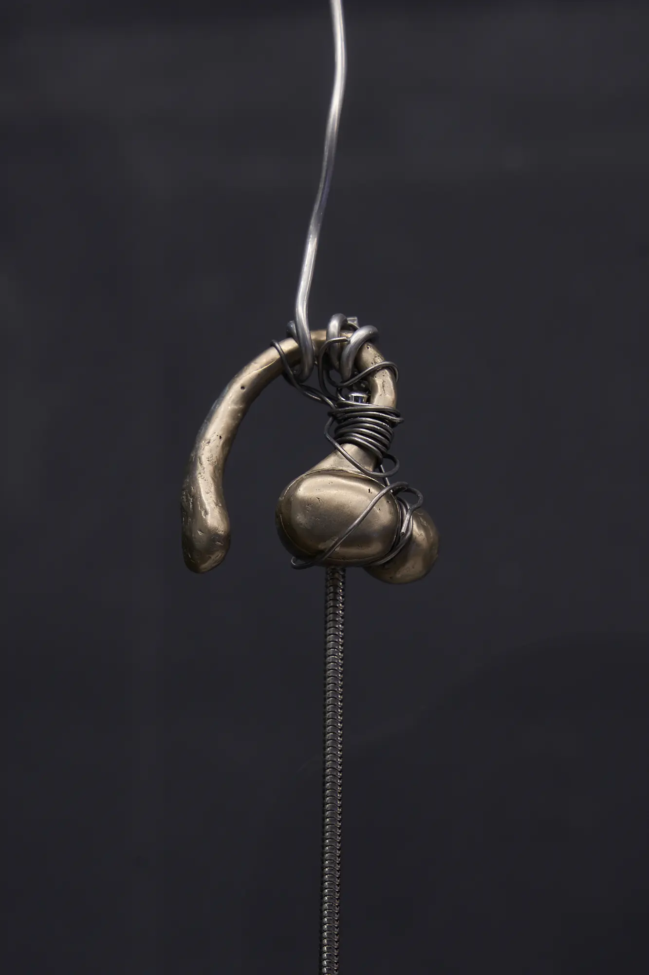 A close-up of a bronze-toned sculptural pendant, wrapped in wire, suspended from a chain against a dark background.


