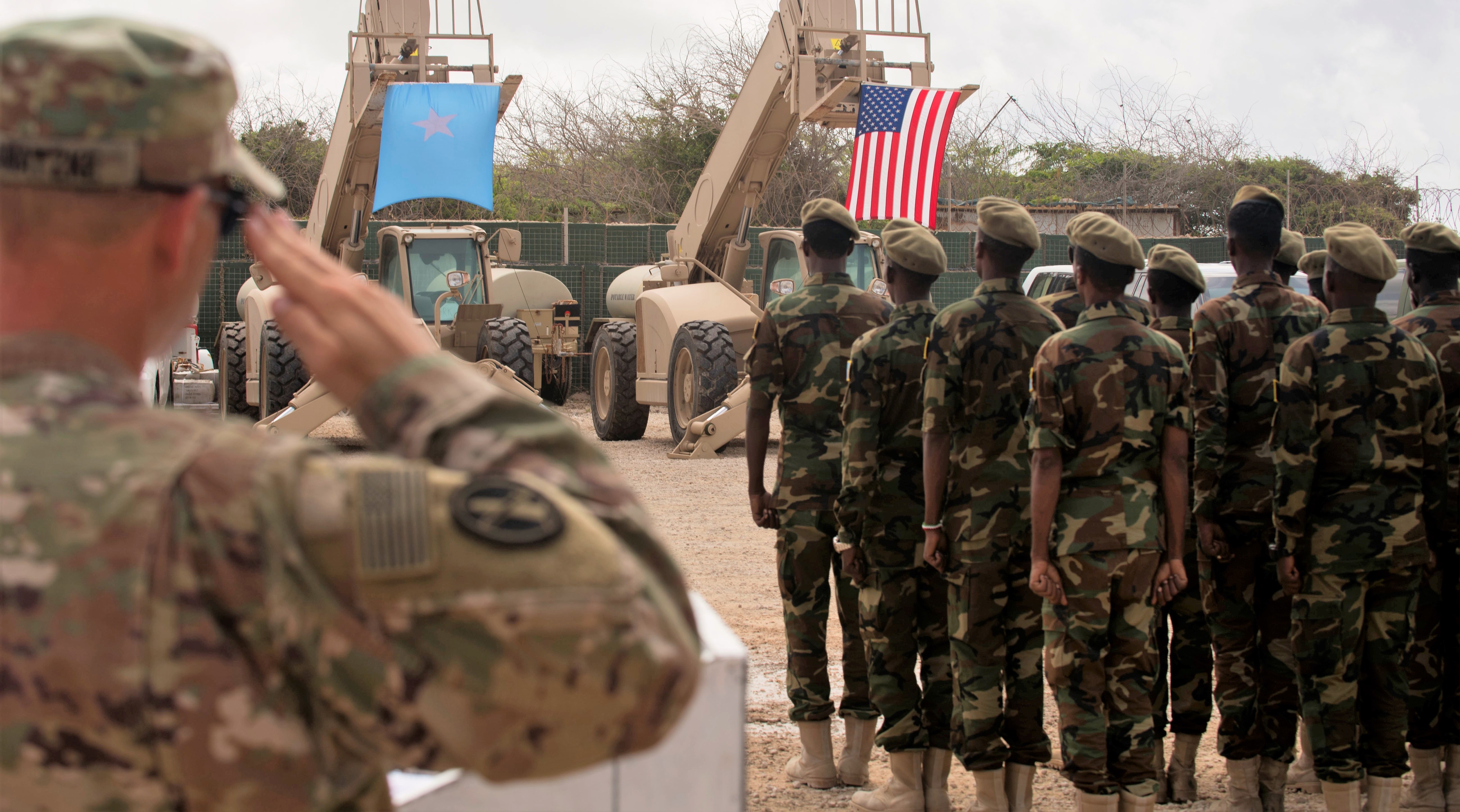 A U.S. soldier salutes as a formation of Somali soldiers stand at attention facing the Somali and U.S. flags, which hang from military vehicles during a joint ceremony in Somalia.