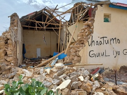 A partially collapsed house in Lasanod with its roof torn away and walls shattered, rubble covering the foreground. Somali graffiti is painted on one remaining wall, while broken timber and masonry fill the exposed interior under a cloudy sky.