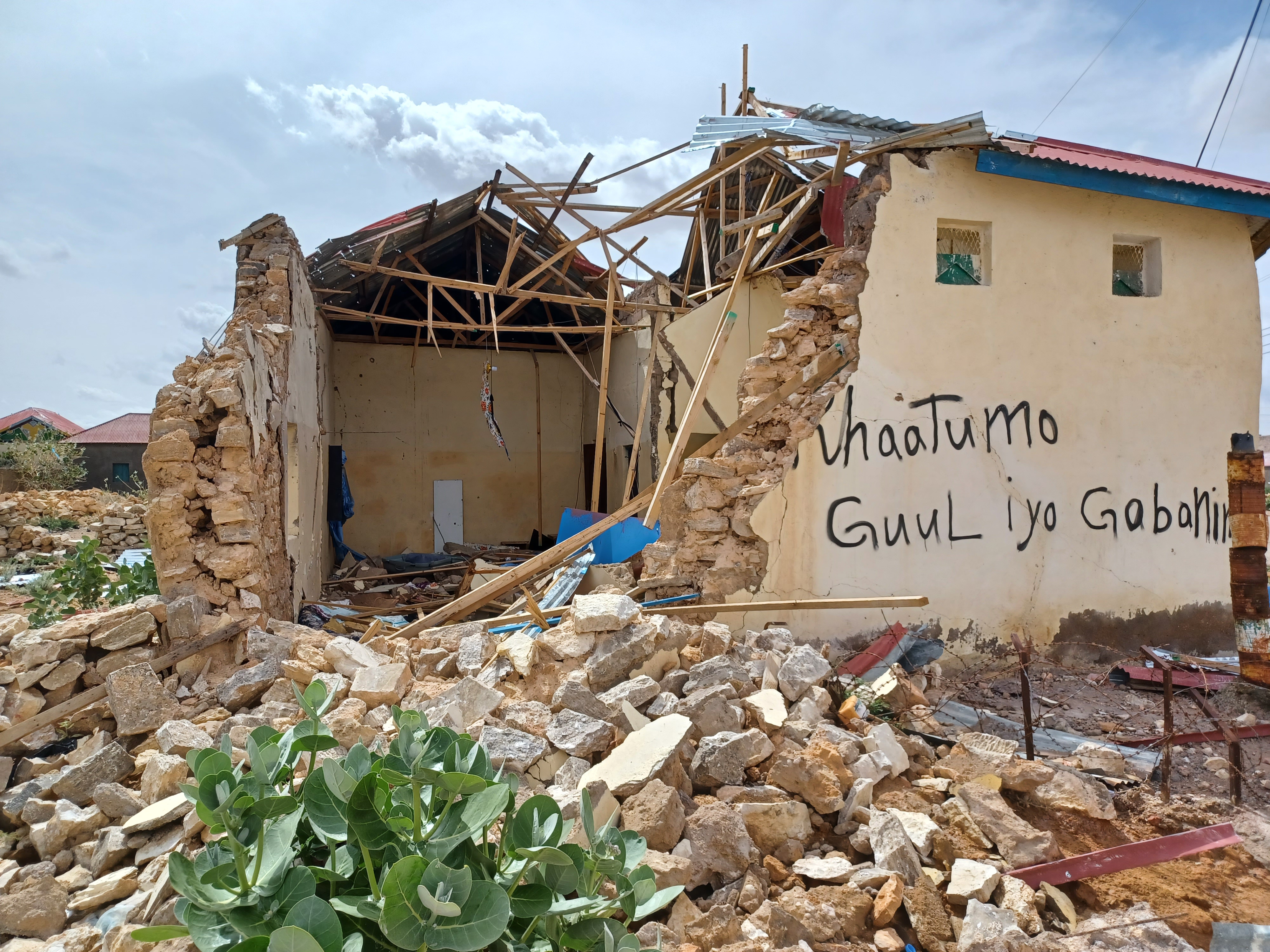 A partially collapsed house in Lasanod with its roof torn away and walls shattered, rubble covering the foreground. Somali graffiti is painted on one remaining wall, while broken timber and masonry fill the exposed interior under a cloudy sky.