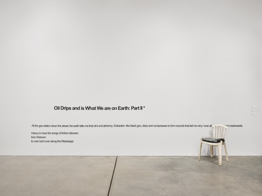 Installation view of a white gallery wall bearing YATTA’s text Oil Drips and is What We are on Earth: Part II. A record player rests on a white plastic chair beside the text.