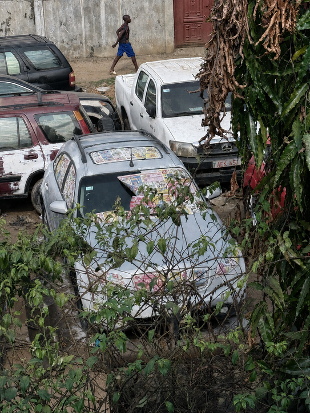 An aerial view of a yard in Freetown filled with parked and disassembled cars, one with faded newspaper plastered across its windshield, as a man walks past in the background.