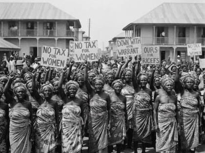  black and white historical photograph from 1929 showing a large crowd of Nigerian women protesting outdoors. They are wearing traditional wrapped clothing and matching headties, with many raising their fists in unison. Several women hold handwritten cardboard signs with slogans including "NO TAX FOR WOMEN", "DOWN WITH WARRANT CHIEFS", and "OURS FOR RICEFS". In the background, two-story colonial-style buildings with corrugated metal roofs stand under an open sky.