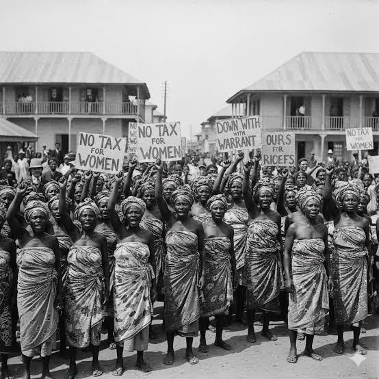  black and white historical photograph from 1929 showing a large crowd of Nigerian women protesting outdoors. They are wearing traditional wrapped clothing and matching headties, with many raising their fists in unison. Several women hold handwritten cardboard signs with slogans including "NO TAX FOR WOMEN", "DOWN WITH WARRANT CHIEFS", and "OURS FOR RICEFS". In the background, two-story colonial-style buildings with corrugated metal roofs stand under an open sky.