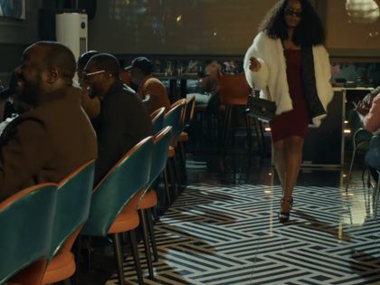 A stylishly dressed woman walks through a busy restaurant as diners watch.
