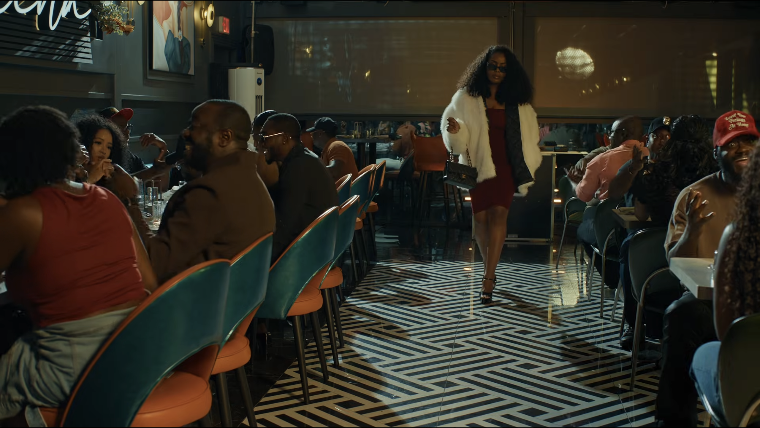 A stylishly dressed woman walks through a busy restaurant as diners watch.