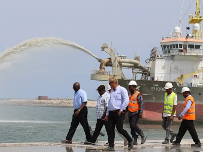 A dredging vessel sprays sand and sediment while workers wearing hard hats walk along newly reclaimed shoreline at the Dangote refinery construction site in Lekki, Lagos State, Nigeria.