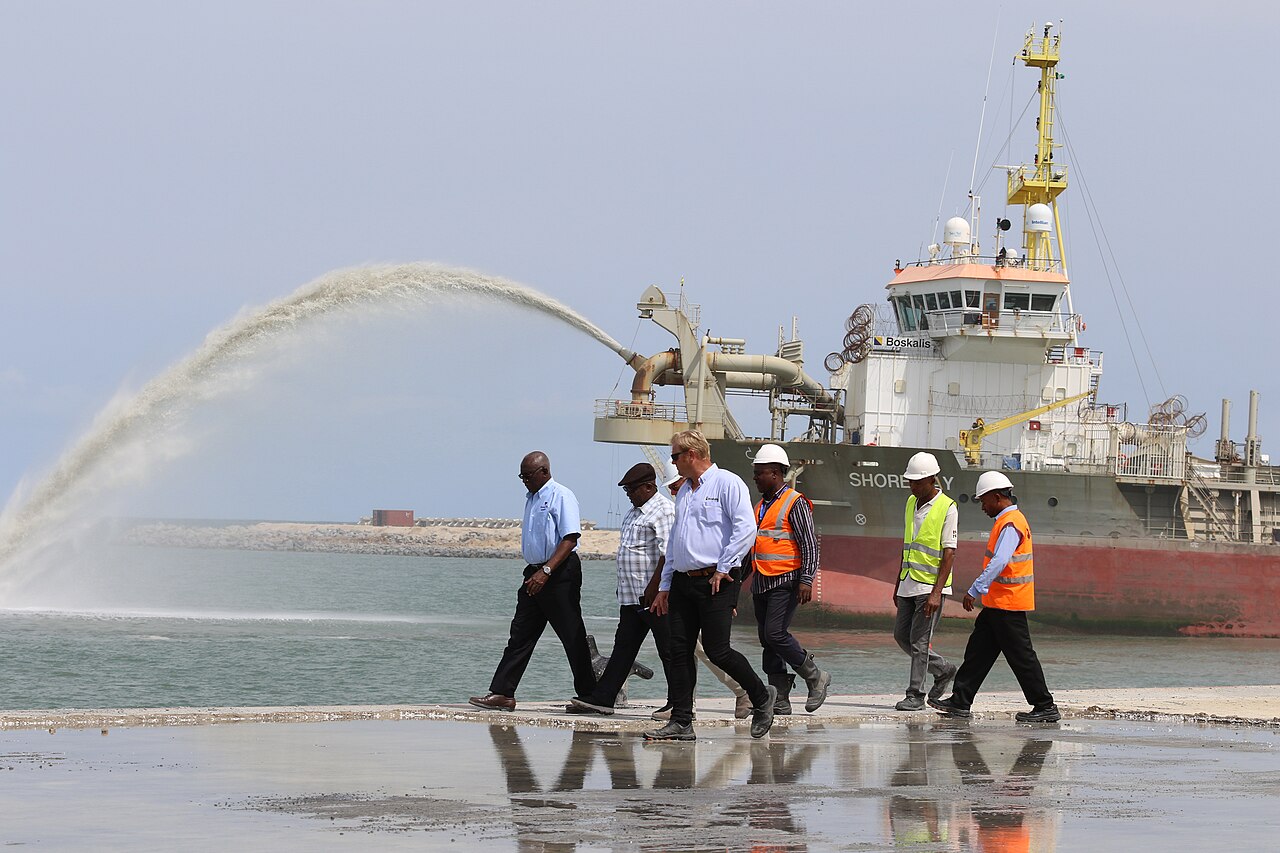 A dredging vessel sprays sand and sediment while workers wearing hard hats walk along newly reclaimed shoreline at the Dangote refinery construction site in Lekki, Lagos State, Nigeria.